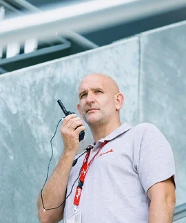 Ein Sicherheitsmitarbeiter spricht in ein Walkie-Talkie, welches er in der rechten Hand an seinen Mund hält. Er trägt ein graues Poloshirt mit einem Klüh-Logo auf der Brust. Im Hintergrund sieht man ein Stück Fassade eines Gebäudes.