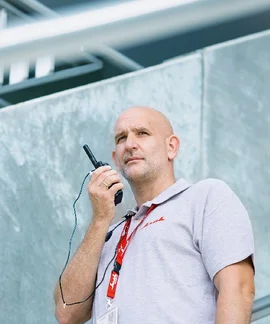 Ein Sicherheitsmitarbeiter spricht in ein Walkie-Talkie, welches er in der rechten Hand an seinen Mund hält. Er trägt ein graues Poloshirt mit einem Klüh-Logo auf der Brust. Im Hintergrund sieht man ein Stück Fassade eines Gebäudes.