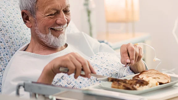 Ein bild von einem Mann im Krankenhausbett und vor sich sein frühstück auf ein Tablett mit einem Glas Orangensaft neben ihm eine Krankenschwester mit einem Klemmbrett und Kugelschreiber