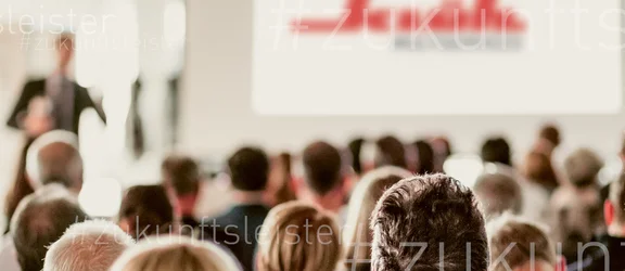 Ein Bild von einem Vortrag von Klüh Multiservices, man sieht viele sitzende Menschen die alle auf die Bühne zum redner schauen