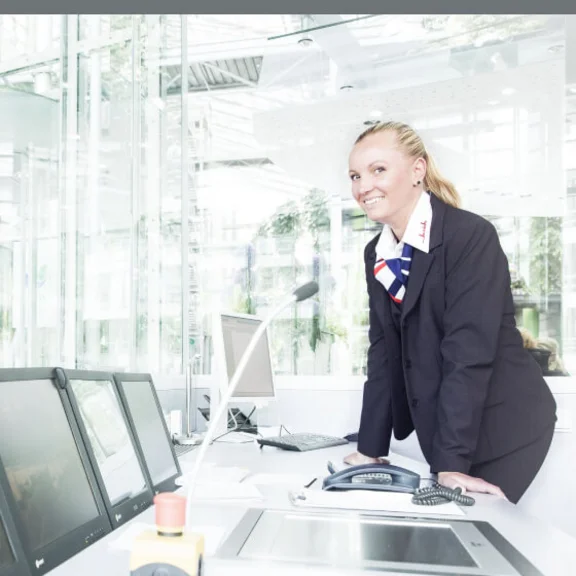 Eine weibliche Servicekraft steht lächelnd an einem Arbeitsplatz mit mehreren Bildschirmen.