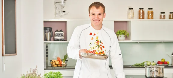 Ein lächelnder Koch in Arbeitskleidung steht in der Küche und schwingt eine Pfanne mit Gemüse drin