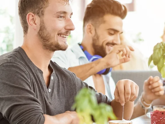 Ein Bannerbild mit Menschen die zusammen am Tisch sitzend essen und trinken. Klüh Catering Logo ist Bild