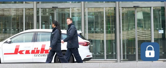Eine weibliche und eine männliche Sicherheitskraft stehen vor einem weißen Auto mit dem Klüh-Logo. Das Auto steht vor dem Eingangsbereich eines großen Bürogebäudes.