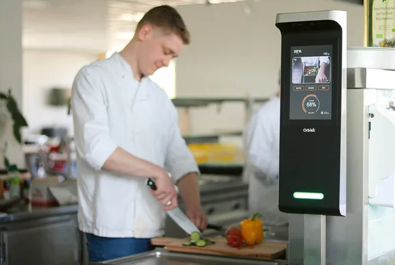 Ein Bild von einem Koch der in einer Küche steht und Gemüse auf einem Schneidebrett schneidet und neben ihm ist ein Orbisk Lebensmittelabfall Gerät