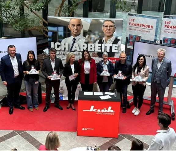 Ein Gruppenbild von neun lächelnden Menschen die nebeneinander stehen auf einem roten Teppich, vor ihnen ist ein Pult von Klüh Multiservices und sie schauen auf ein Publikum vor sich