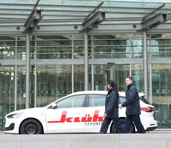 Ein Bild von einem Mann und eine Frau in Klüh Security Arbeitskleidung gehen eine Straße entlang und neben ihnen ein Klüh Security Firmenauto