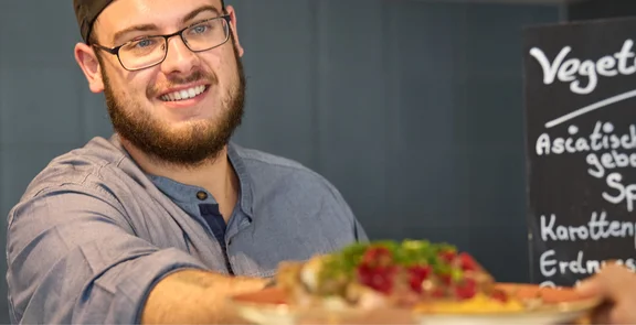 Ein Koch übergibt ein Tellergericht, im Hintergrund ist eine Wand mit dunkelgrauen Fließen und ein Kreidetafel mit der Aufschrift " Vegetarisch Asiatisch gebackene Spitzkohl, Karottenpolenta, Erdnusssauce
