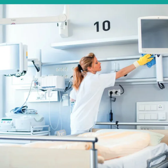 Eine weibliche Reinigungskraft wischt in einem Krankenhauszimmer Oberflächen ab.
