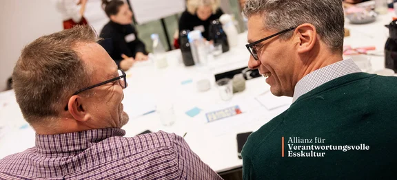 Ein Bild von zwei Männern die an einem Besprechungstisch sitzen und sich lächelnd angucken, im Hintergrund sieht man weitere Personen am Tisch sitzen, am Rand des Bildes ist der Slogan "Allianz für Verantwortungsvolle Esskultur"