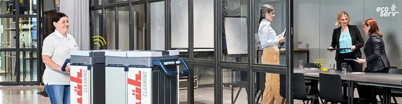 Ein Bild von einer Klüh Cleaning Reinigungskraft meinem Klüh Cleaning Reinigungswagen im Flur eines Bürogebäudes, im Hintergrund sind drei stehende Frau in einem Besprechungsraum mit einer Glaswand