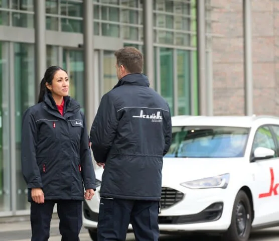 Ein Bild von einer Frau und einem Mann in Klüh Security Arbeitskleidung stehen sich gegenüber und im Hintergrund ist ein Klüh Security Firmenauto