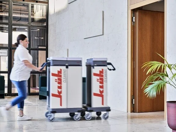 Ein Bild von einer Frau die ein Klüh Cleaning Reinigungswagen durch ein Flur schiebt