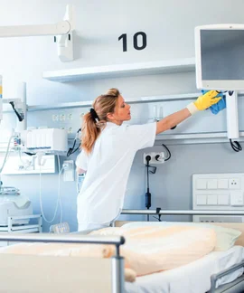 Eine Frau in weißer Kleidung reinigt die Ablage oberhalb eines Krankenhausbettes. Im Hintergrund sind Bildschirme und medizinische Technik zu erkennen.