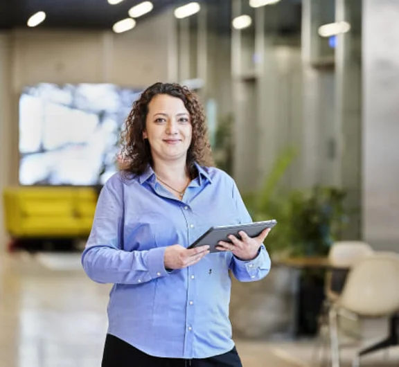 eine Frau die lächelnd in die Kamera schaut und hält dabei ein Tablet in ihren Händen