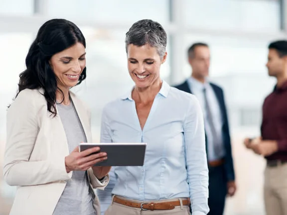 Ein Bild von Frauen die lächelnd nebeneinander stehen und auf ein Tablet schauen, im Hintergrund sind zwei Männer die sich gegenüberstehen und anschauen